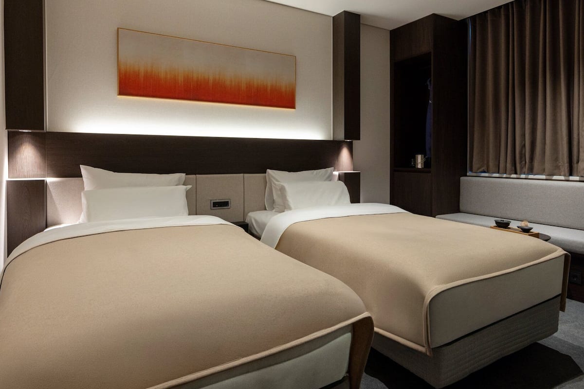 A hotel room with two beds, a desk, a chair, a large painting, and a window with curtains.