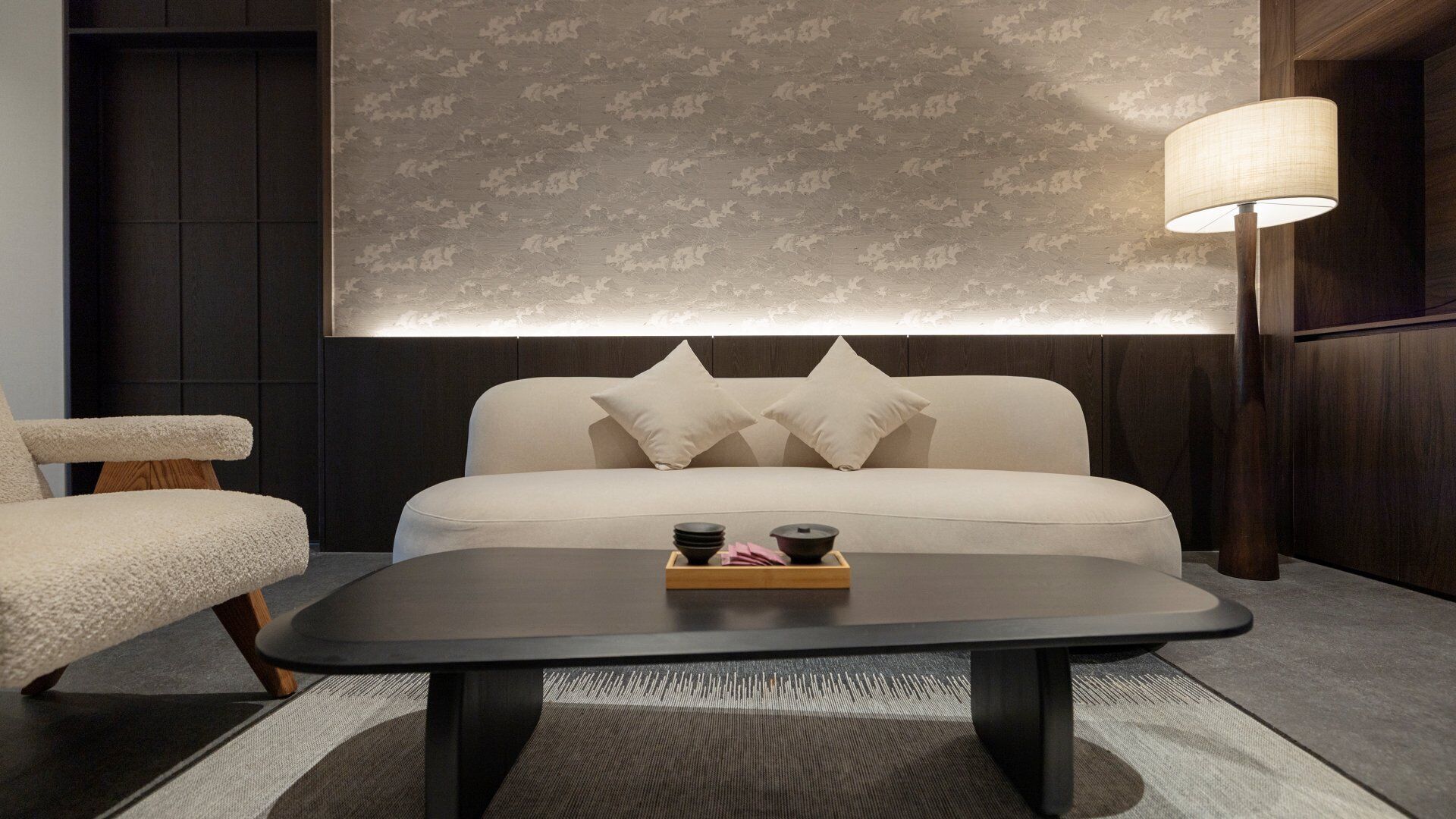 A modern hotel room with a sofa, a coffee table with a tray of items, and a lamp.