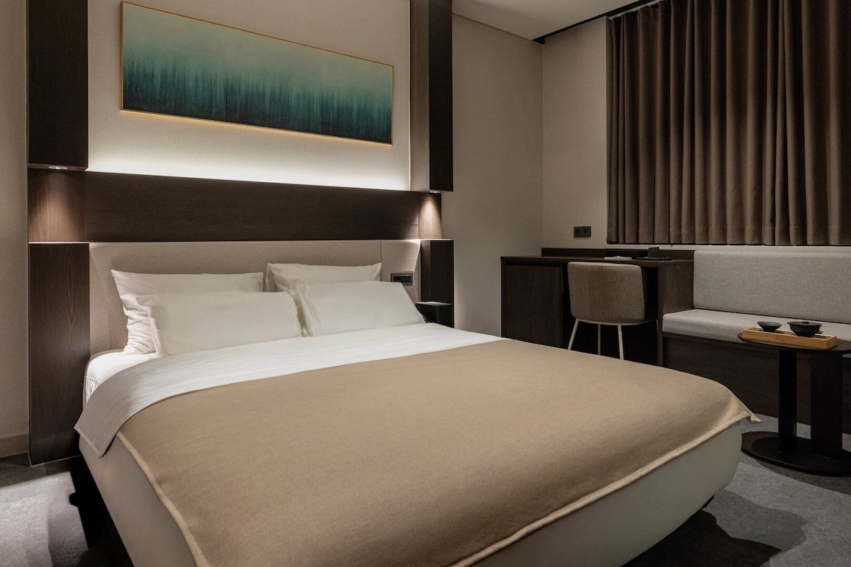 A modern hotel room with a large bed, a desk, a chair, and a painting on the wall.