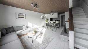 A modern living room with a sofa, coffee table, and dining area. There is a staircase leading to an upper level. - HILLS NISEKO E (Niseko)
