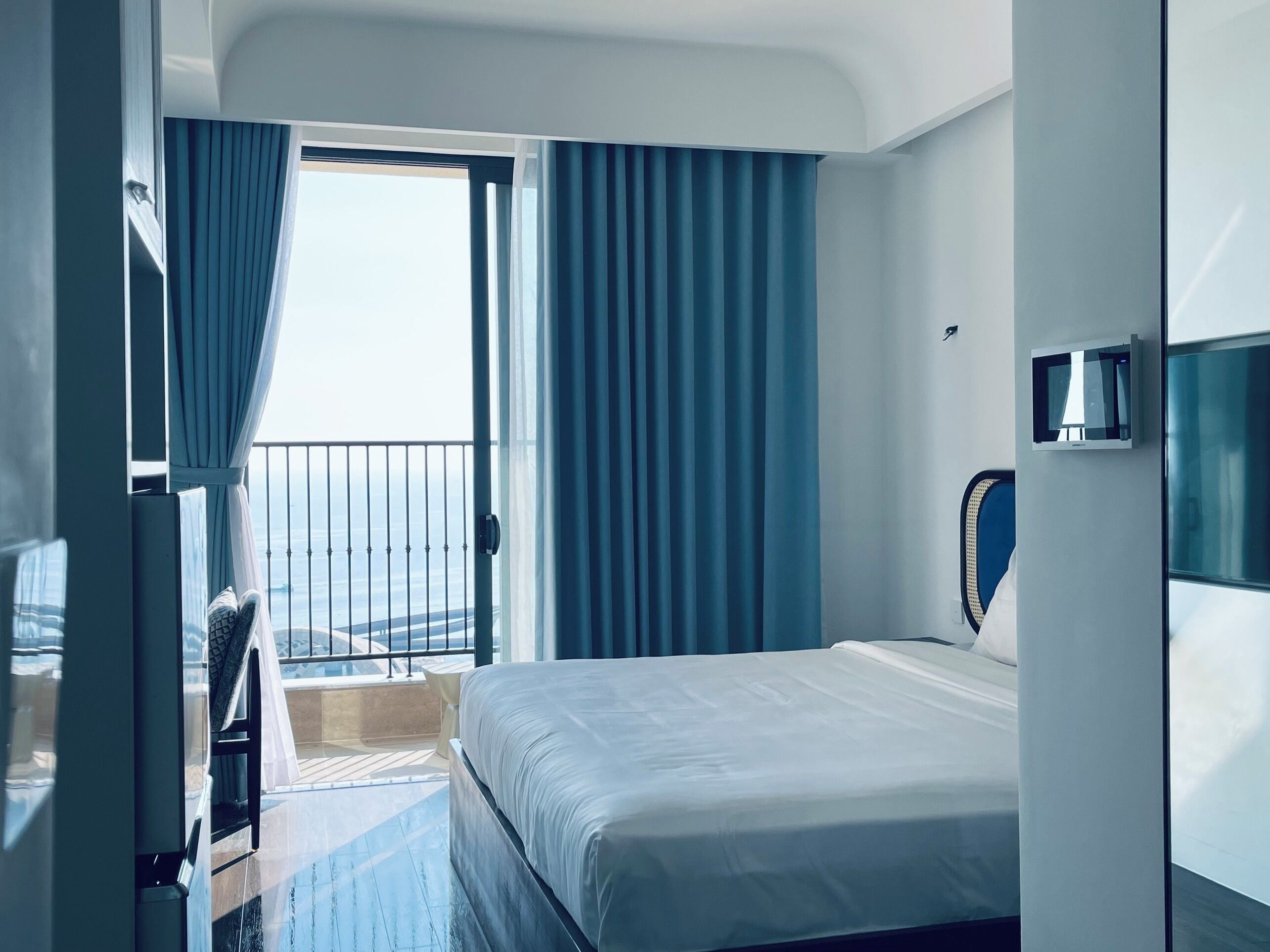 Une chambre d’hôtel moderne avec un grand lit, un téléviseur fixé au mur et un balcon agrémenté de rideaux bleus.