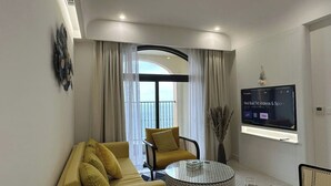 Una sala de estar moderna con un sofá, un sillón y una mesa de centro redonda, un televisor de pantalla plana montado en la pared y un balcón con cortinas.
