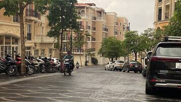 Une rue mouillée, bordée de voitures et de motos garées, des immeubles avec des balcons et un trottoir bordé d’arbres.