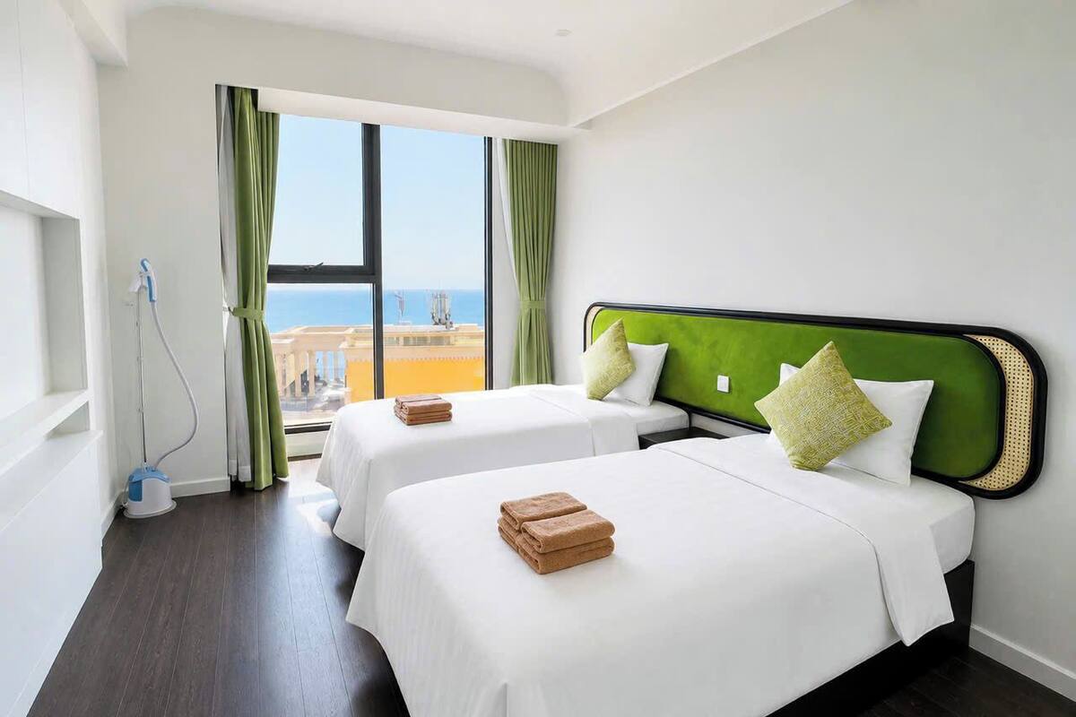 Habitación de hotel con dos camas, un ventanal con cortinas y vista al mar, además de un aspirador.