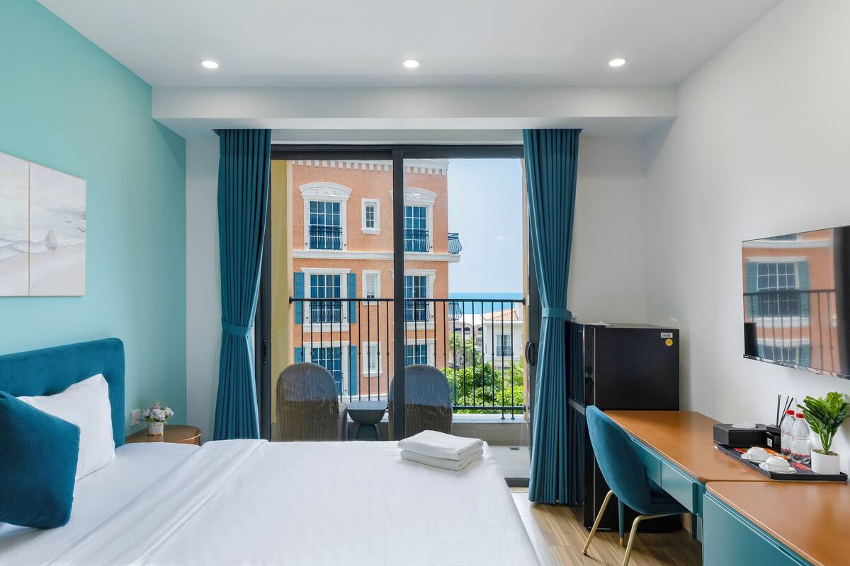 Habitación de hotel con cama, escritorio, silla, televisor y balcón con vistas a edificios.