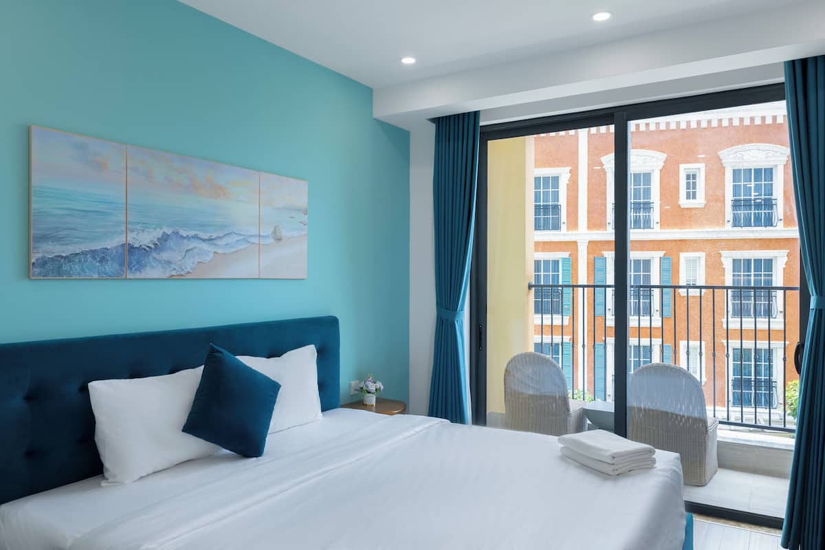 Habitación de hotel con cama, un cuadro, vista de un edificio a través de una ventana y un balcón con sillas de mimbre.