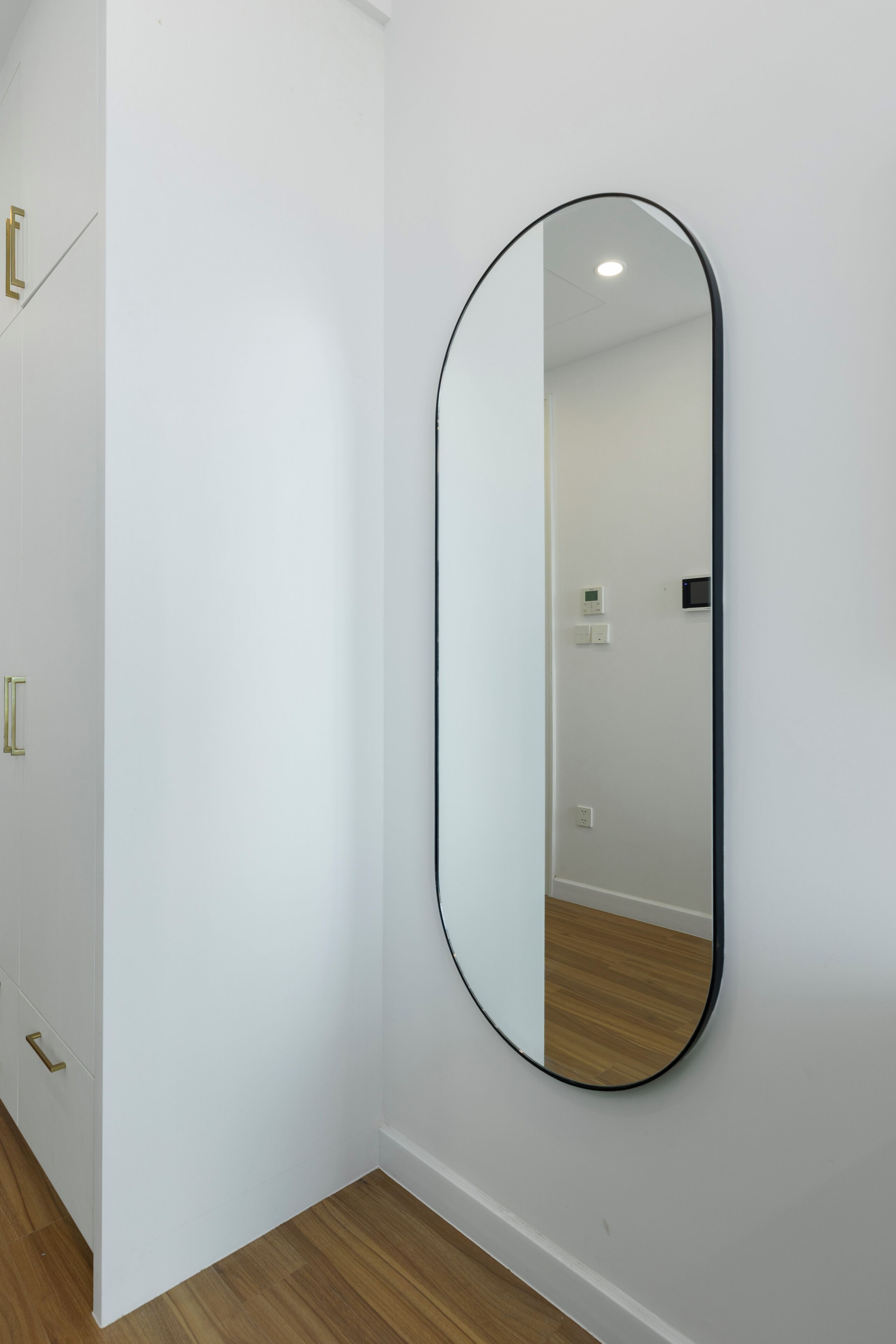 A room with a white wall, a wooden floor, and a large oval mirror.