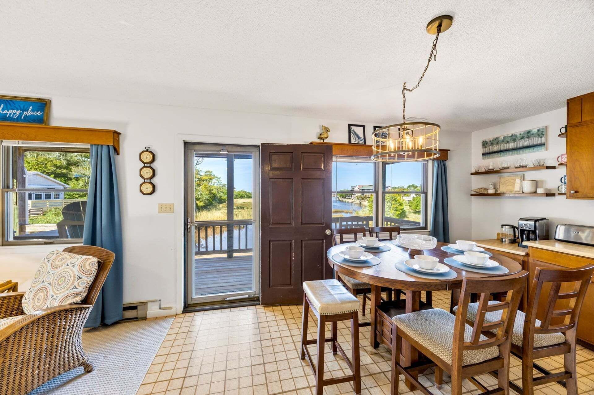 A dining area with a view of the outdoors, a wooden table set for four, and a kitchen area with a coffee maker and shelves.