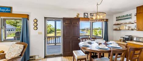A dining area with a view of the outdoors, a wooden table set for four, and a kitchen area with a coffee maker and shelves.
