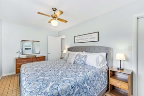 A bedroom with a bed, a nightstand, a lamp, a ceiling fan, a mirror, and a framed sign on the wall.