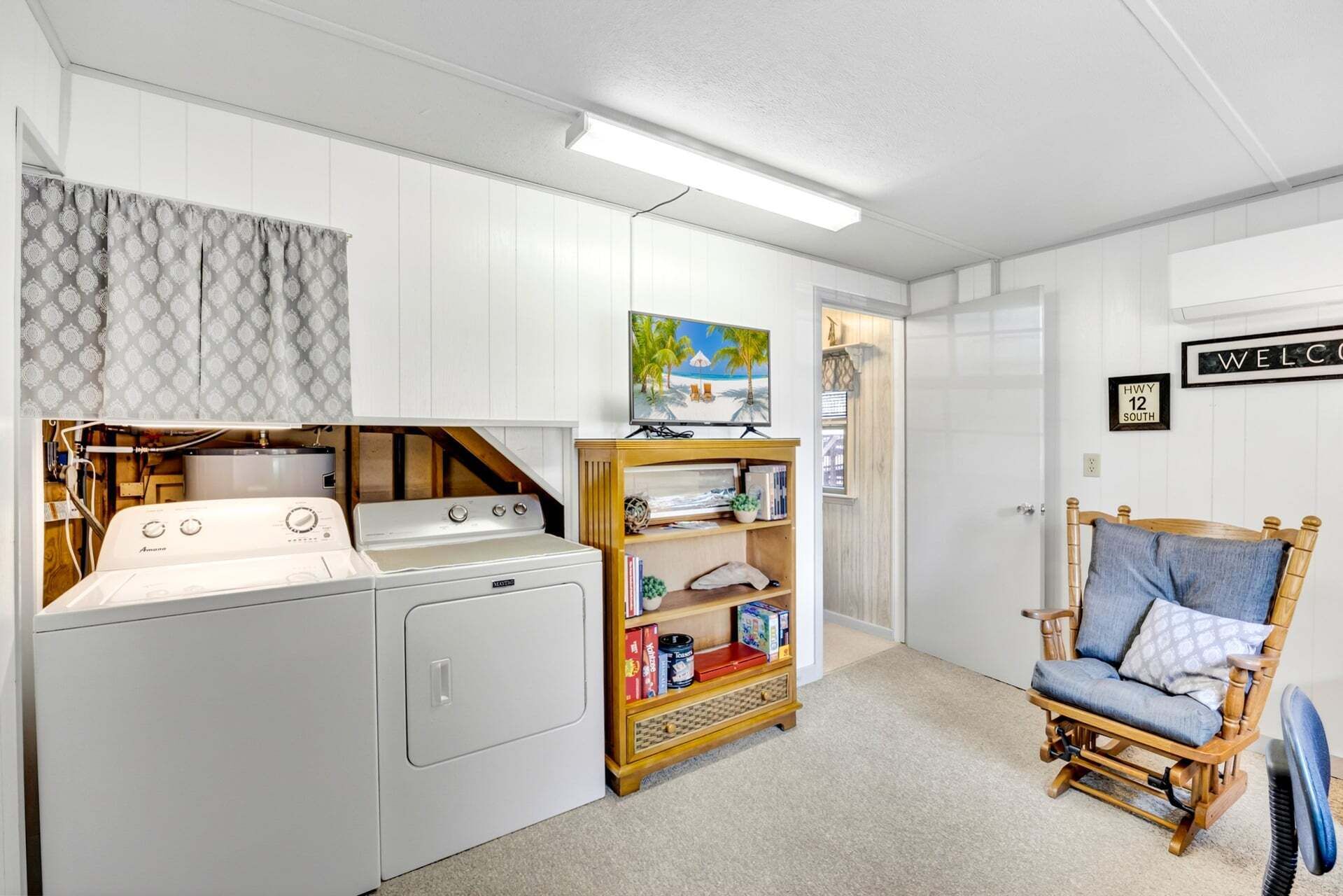 Un espace buanderie avec un lave-linge et un sèche-linge, une étagère en bois avec des livres et des jeux, une chaise en osier et un tableau représentant une scène de plage.