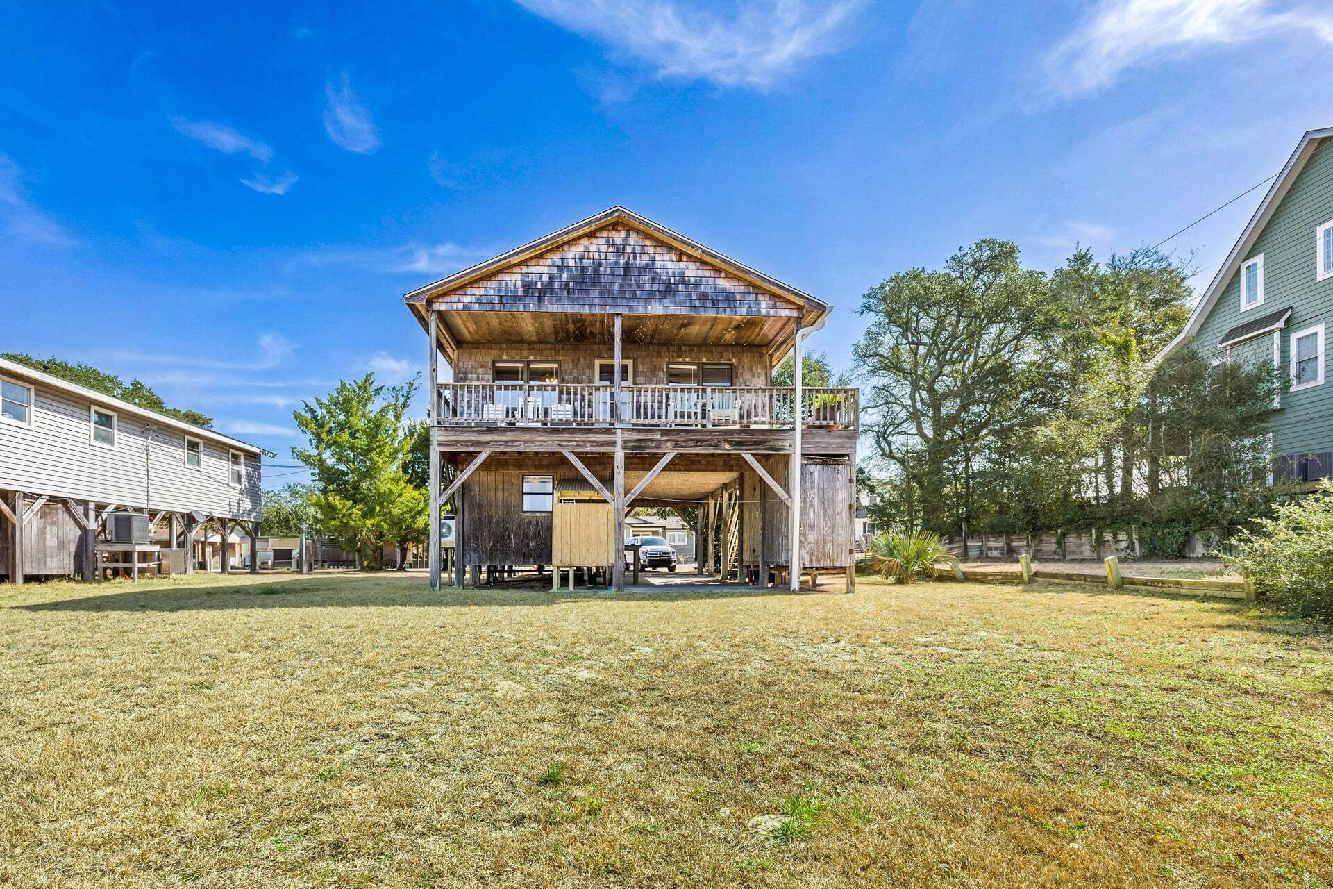 Une maison en bois à deux étages, dotée d’un balcon, entourée d’une cour verdoyante et d’arbres.