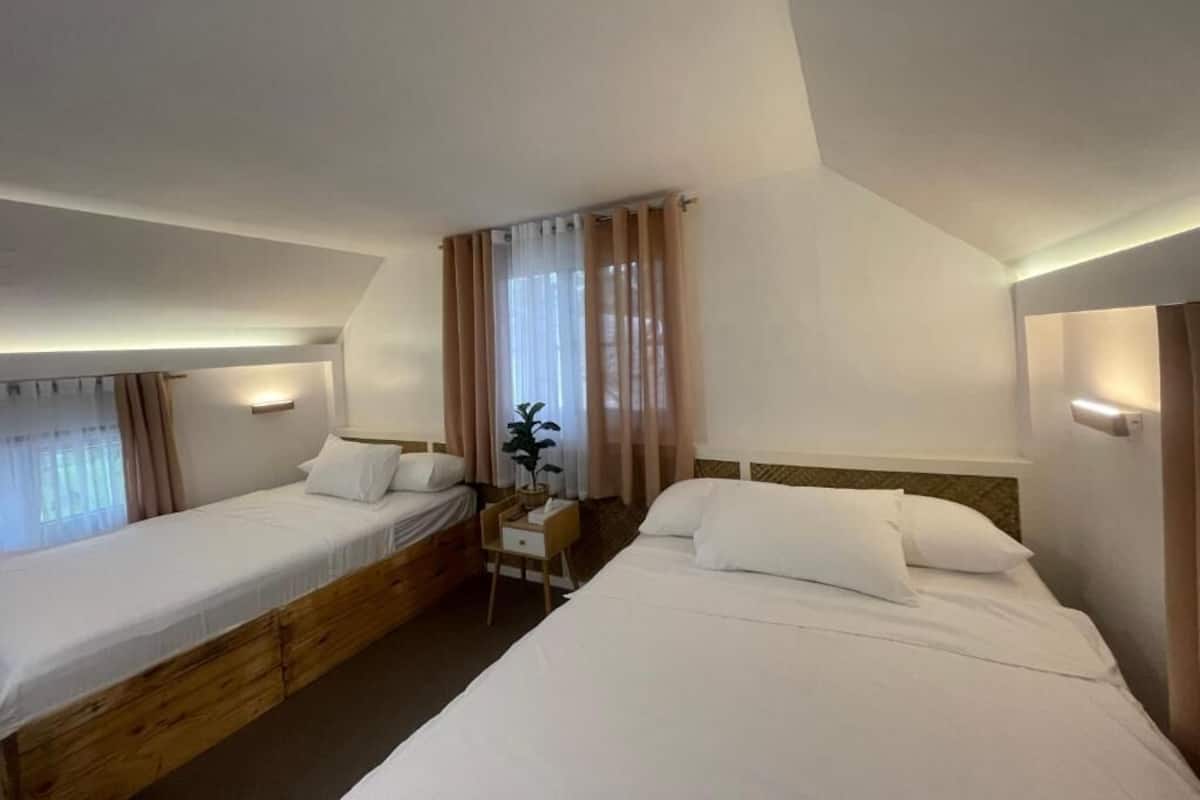 A hotel room with two single beds, a small bedside table with a lamp, and a window with curtains.