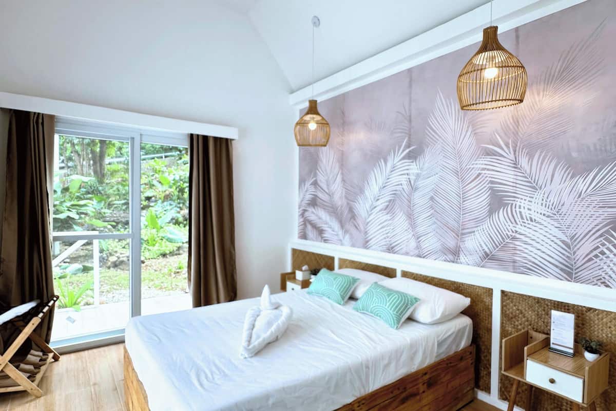 A bedroom with a wooden bed, a ceiling fan, a large window with curtains, a wall-mounted light fixture, and a patterned wall.