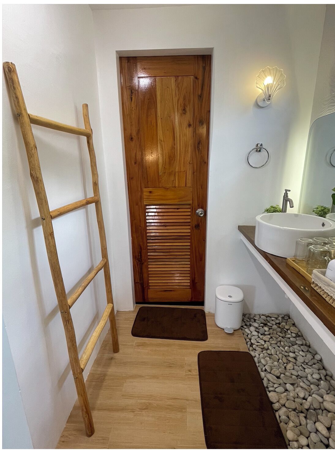 Una puerta de madera con un picaporte metálico, un lavabo blanco, un espejo y un pequeño cubo de basura blanco.