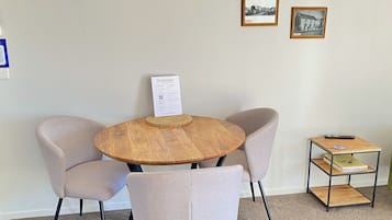 A round wooden table with a welcome sign, two upholstered chairs, and a small metal shelf with books and a remote control.