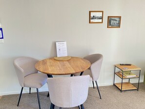 A round wooden table with a welcome sign, two upholstered chairs, and a small metal shelf with books and a remote control. - Pioneer Motel (Collingwood)