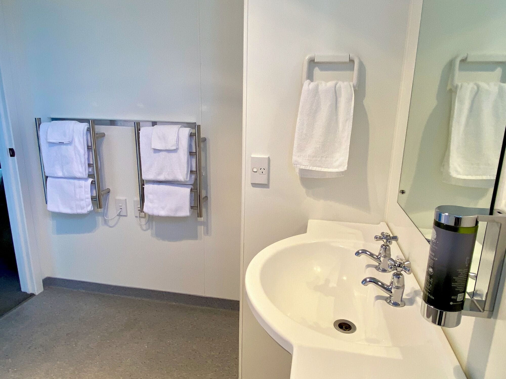 Ein Badezimmer mit einem weißen Waschbecken, zwei Wasserhähnen, einem Spiegel und einem Handtuchhalter mit mehreren Handtüchern.