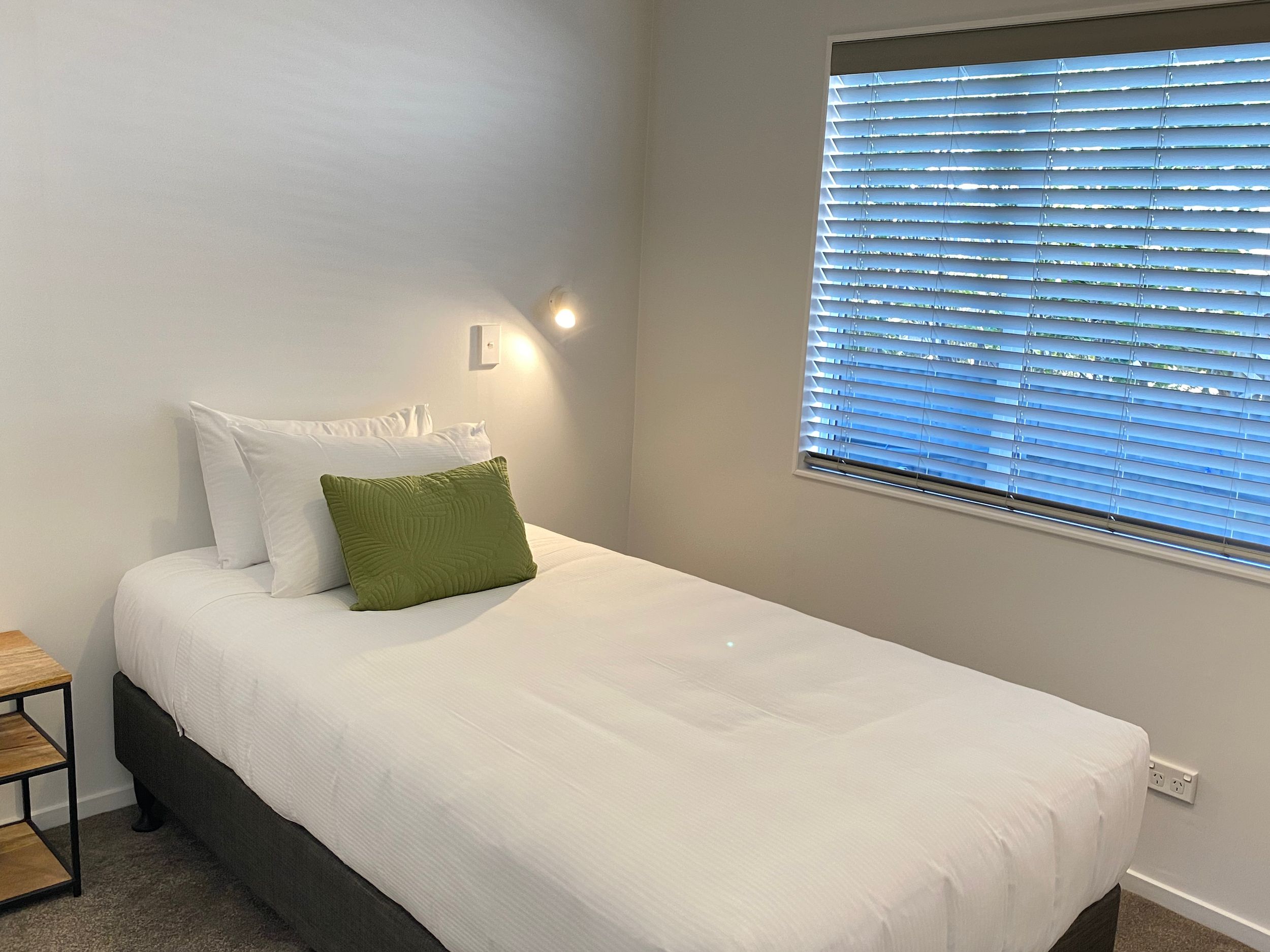 Ein Einzelbett mit weißer Bettwäsche und einem grünen Kopfkissen, ein Nachttisch mit einer Lampe und ein Fenster mit Jalousien.