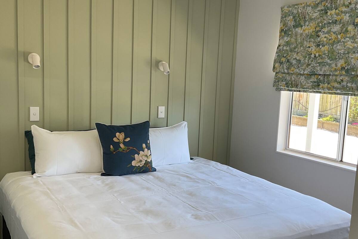 A bed with white bedding and a blue floral pillow, a green wall with vertical panels, and a window with a floral blind.