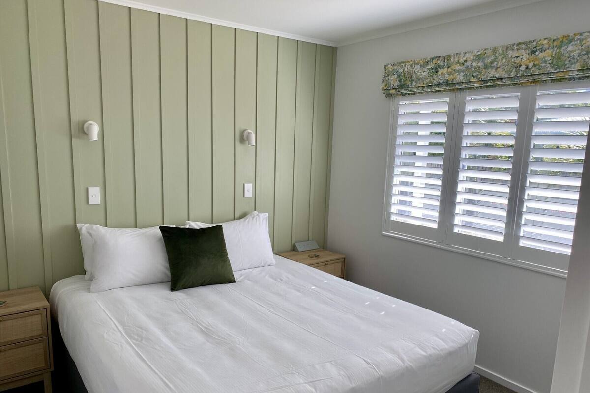 A neatly made bed with white and dark green pillows, a wooden nightstand with a lamp, and a window with blinds.
