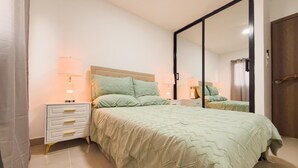 Een slaapkamer met een groot bed, een nachtkastje met een lamp, een dressoir en een spiegel.