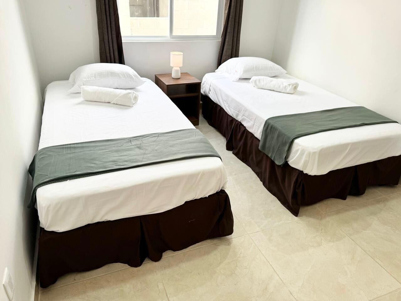 A room with two single beds, each with a white mattress and dark skirted beddings. There is a small bedside table with a lamp between the beds.
