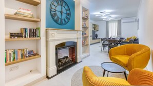 Een woonkamer met een open haard, boekenkasten, een grote klok en een gele fauteuil.