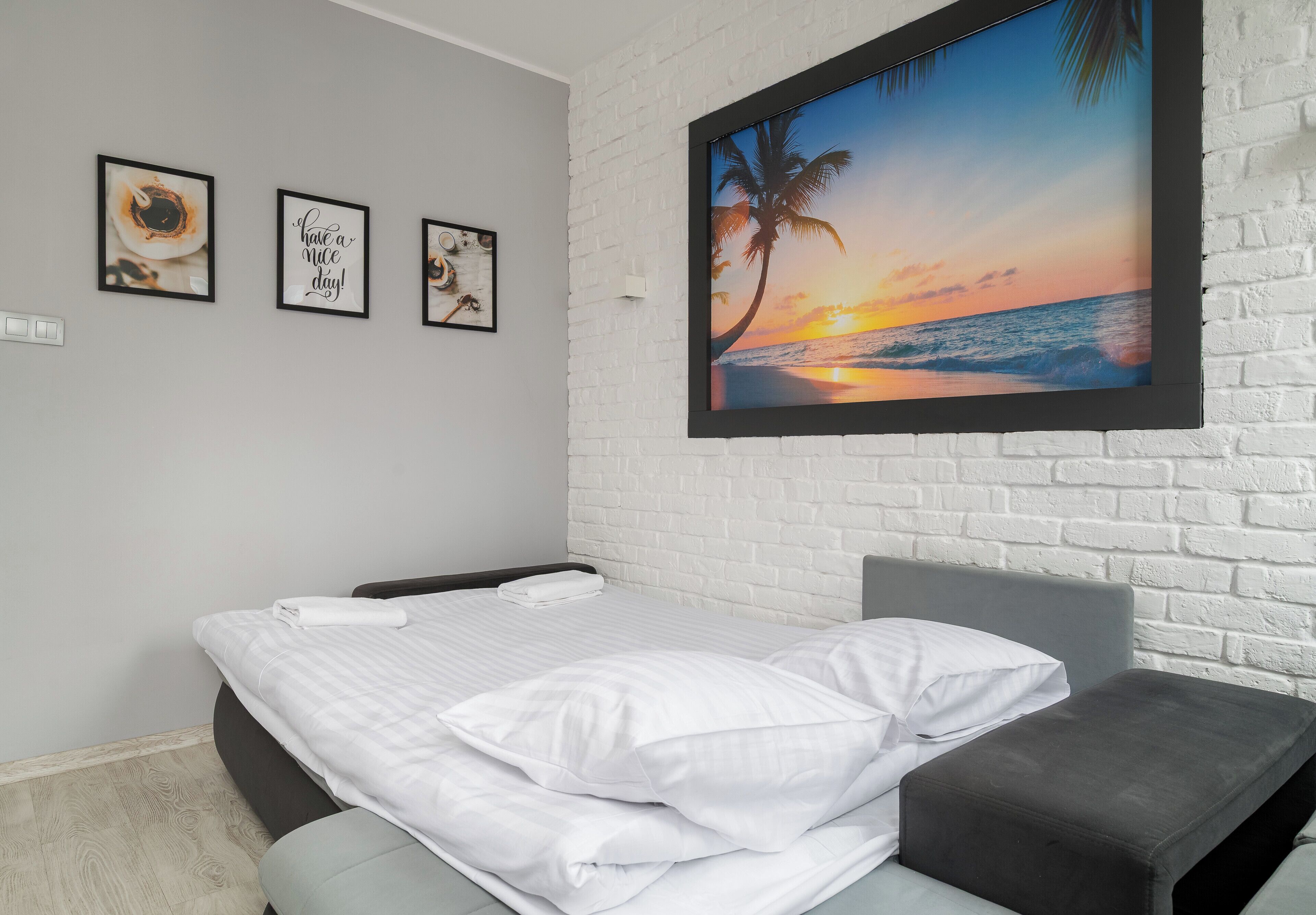 A neatly made bed with white linens, a gray headboard, and a gray bench. There are three framed pictures on the wall, one of which says "have a nice day!" The wall is white brick, and there's a large framed picture of a sunset over the ocean.