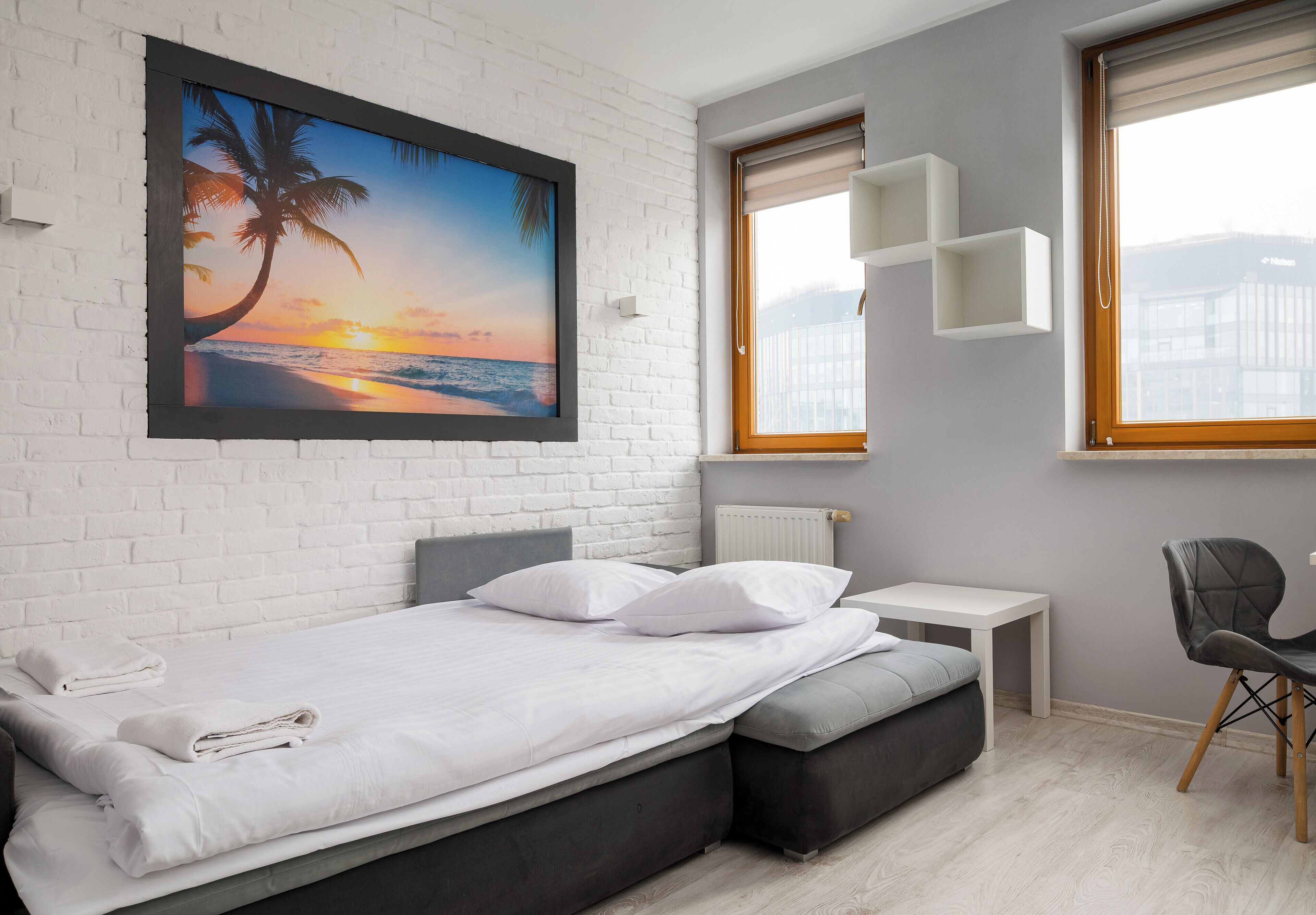 Un dormitorio moderno con pared de ladrillo blanco, una cama con ropa de cama blanca, una mesita de noche pequeña, una silla y un ventanal con vistas al mar.