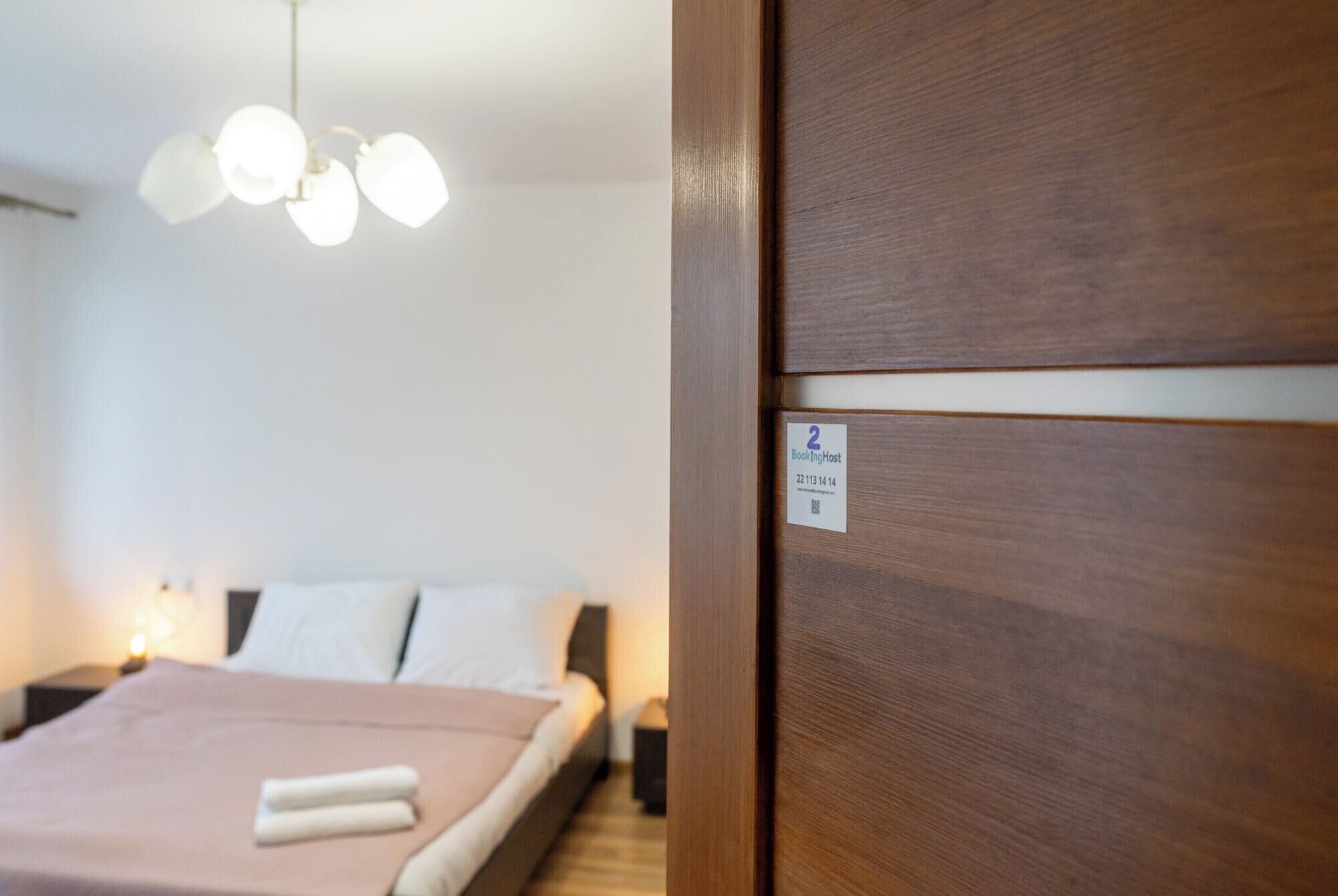 Hotellihuone, jossa on puinen ovi, valkoisilla lakanoiden peitetty sänky ja katosta roikkuva valaisin.