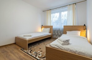 Une chambre avec deux lits simples, une tête de lit en bois, une fenêtre avec des rideaux et un tapis à motifs.