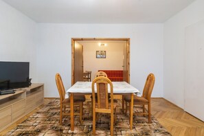 Un coin repas avec une table dressée pour quatre personnes, des chaises en bois, un téléviseur posé sur une étagère et un tapis à motifs.