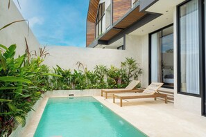 A modern outdoor pool area with two lounge chairs, surrounded by lush greenery and a contemporary building with large windows. - Villa Fresko 5 by Alfred in Bali (Munggu)