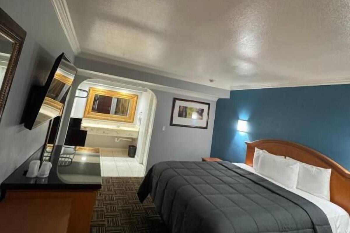 Habitación de hotel con cama, escritorio, espejo, baño con lavamanos y espejo, y un cuadro en la pared.