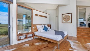 A modern bedroom with a large bed, a glass shower enclosure, wooden flooring, and a view of the outdoors. - Fisherman's Rest - Your Perfect Couple’s Retreat (Port Fairy)