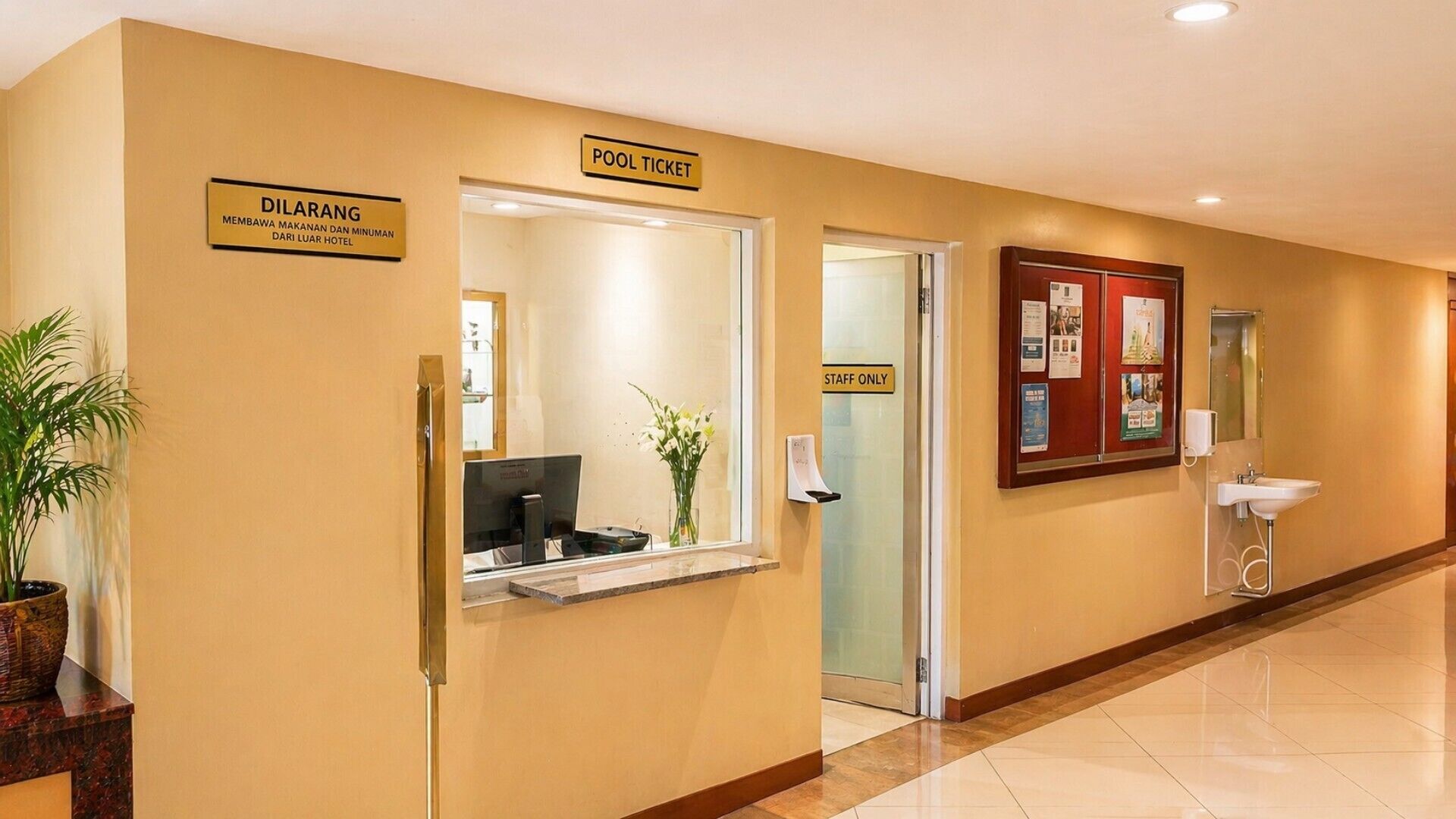 Una zona de recepción con mostrador, un cartel que dice 'POOL TICKET', un cartel que dice 'STAFF ONLY' y un cartel que dice 'DILARANG' que indica prohibido el acceso.
