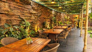 Un patio al aire libre con paredes de piedra, mesas de madera, sillas de mimbre y plantas colgantes.