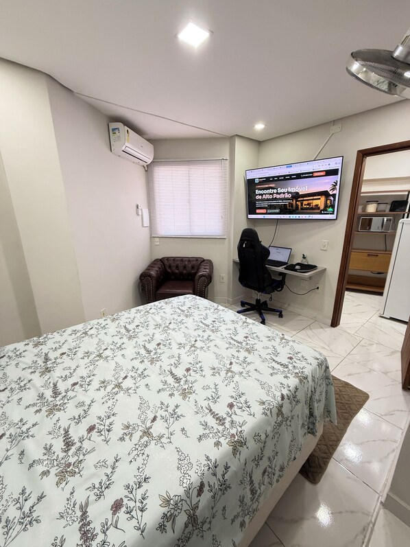 A bedroom with a bed, a desk with a computer, a chair, a television, and a window with blinds. - Full suite with air conditioning, Wi-Fi, and Netflix. (Vitória)