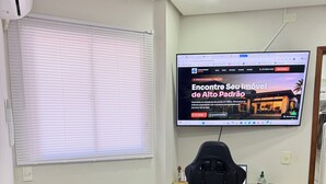 Et værelse med en brun lædersofa, en sort kontorstol, et skrivebord med en computer og et fjernsyn, der viser et ejendomssite.