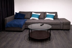 Un canapé gris agrémenté de coussins bleus et blancs, un plaid bleu et une table basse en verre sur laquelle repose une télécommande.
