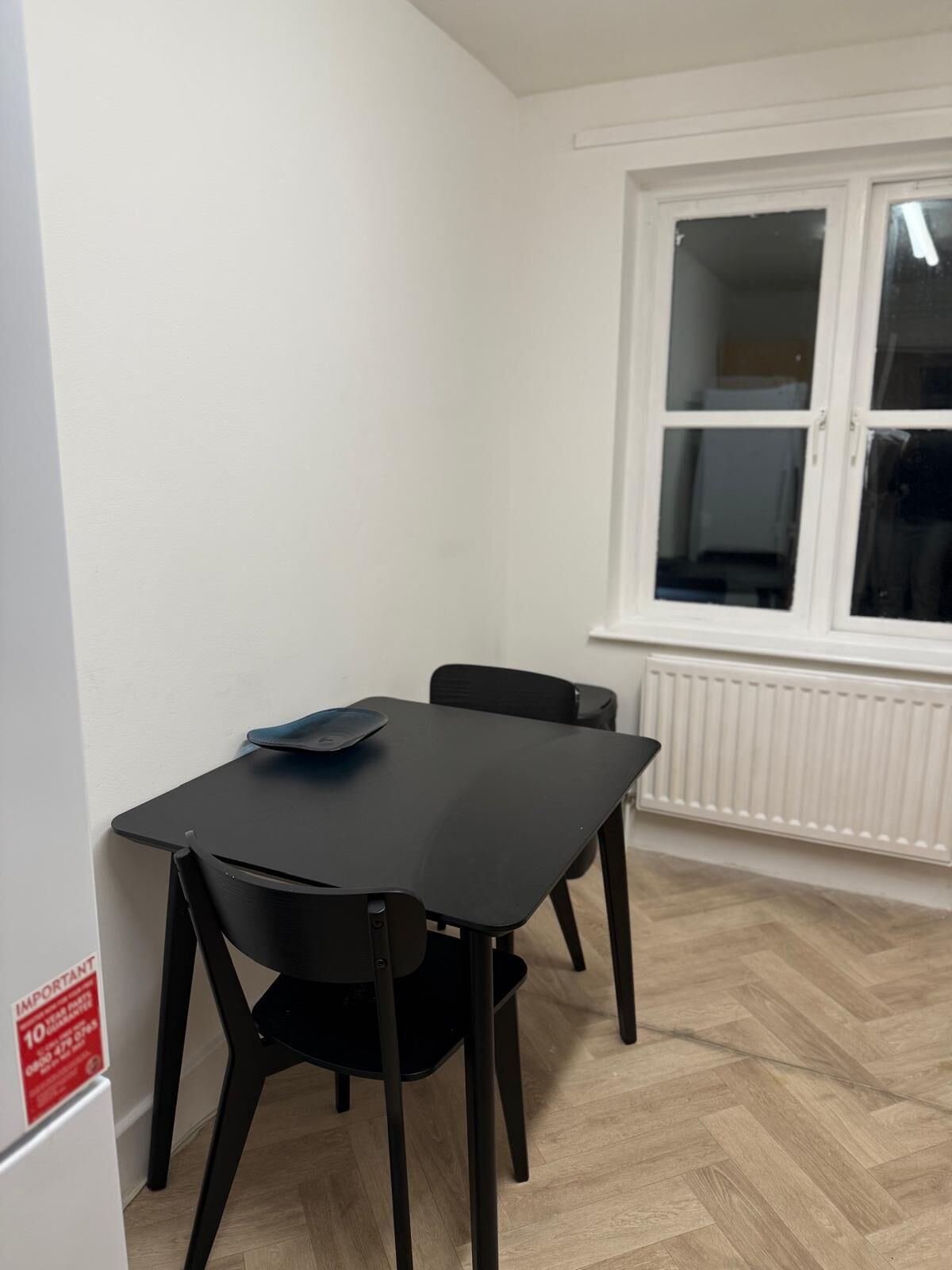 Ein kleiner Raum mit einem schwarzen Tisch, zwei Stühlen, einem weißen Heizkörper und einem Fenster mit Jalousien.
