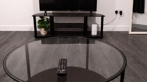 A modern living room with a glass coffee table, a remote control, a flat-screen TV, a black entertainment unit, and a potted plant. - Wonderful 1-bed apt East London central (London)