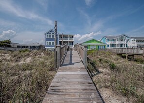 A wooden boardwalk leading to a beachfront property with multiple houses, a wooden fence, and a clear sky. - Carolina Memories - Stunning oceanfront home with private pool and more! (Holden Beach)