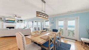 Amplia sala de estar con un gran comedor de madera, sillas tapizadas en azul y vista al océano a través de puertas correderas de vidrio.