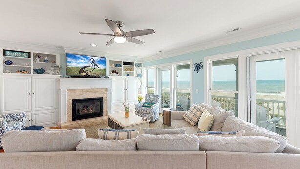 Una sala de estar con chimenea, un sofá grande, un ventilador de techo y vista a la playa.
