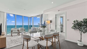 A modern hotel room with a large glass table, grey upholstered chairs, a potted plant, and a view of the ocean. - Hilton 2BR Retreat Sky High Ocean Views Pool (Surfers Paradise)