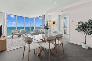 A modern hotel room with a large glass table, grey upholstered chairs, a potted plant, and a view of the ocean. - Hilton 2BR Retreat Sky High Ocean Views Pool (Surfers Paradise)