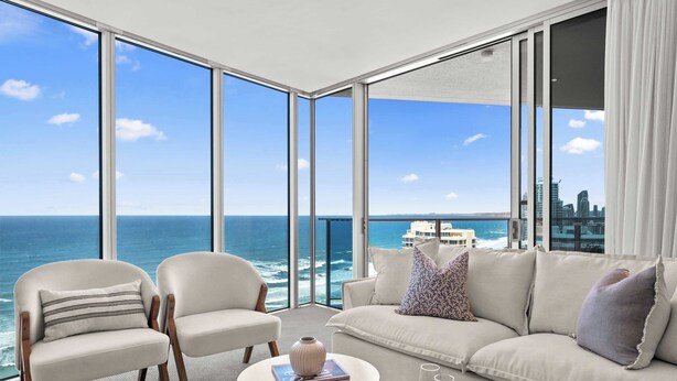 A modern living room with a large window overlooking the ocean, a sofa, two armchairs, and a coffee table with a book and glasses. - Hilton 2BR Retreat Sky High Ocean Views Pool (Surfers Paradise)