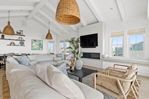 Ein modernes Wohnzimmer mit Ecksofa, Couchtisch und Kamin. Wicker-Pendelleuchten, ein Flachbildfernseher an der Wand und ein großes Fenster mit Ausblick sind zu sehen.